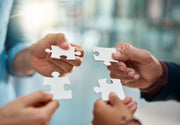 Four hands holding white puzzle pieces against a blurred indoor background