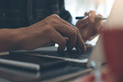 Person typing on a laptop with a smartphone in hand, blurred background