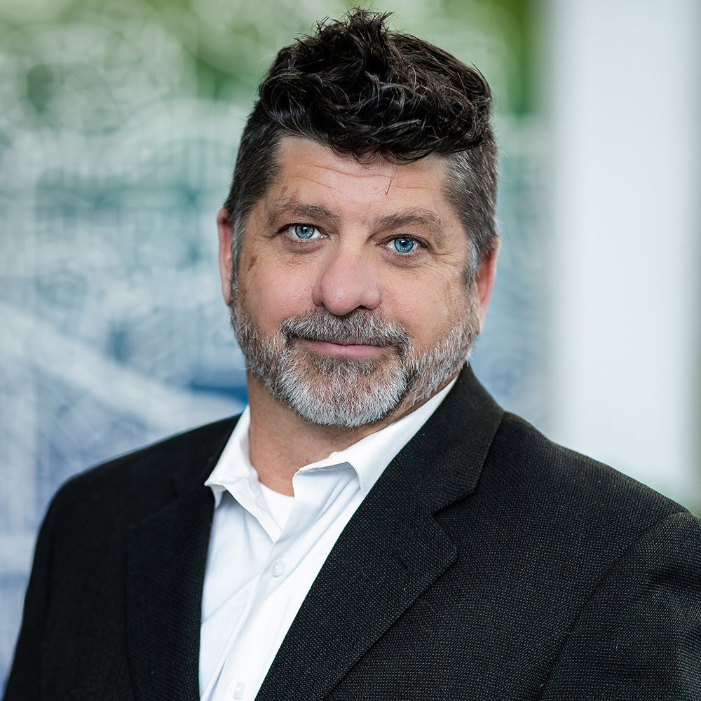 Chris Mapson wearing a black suit and white shirt with a blurred outdoor background