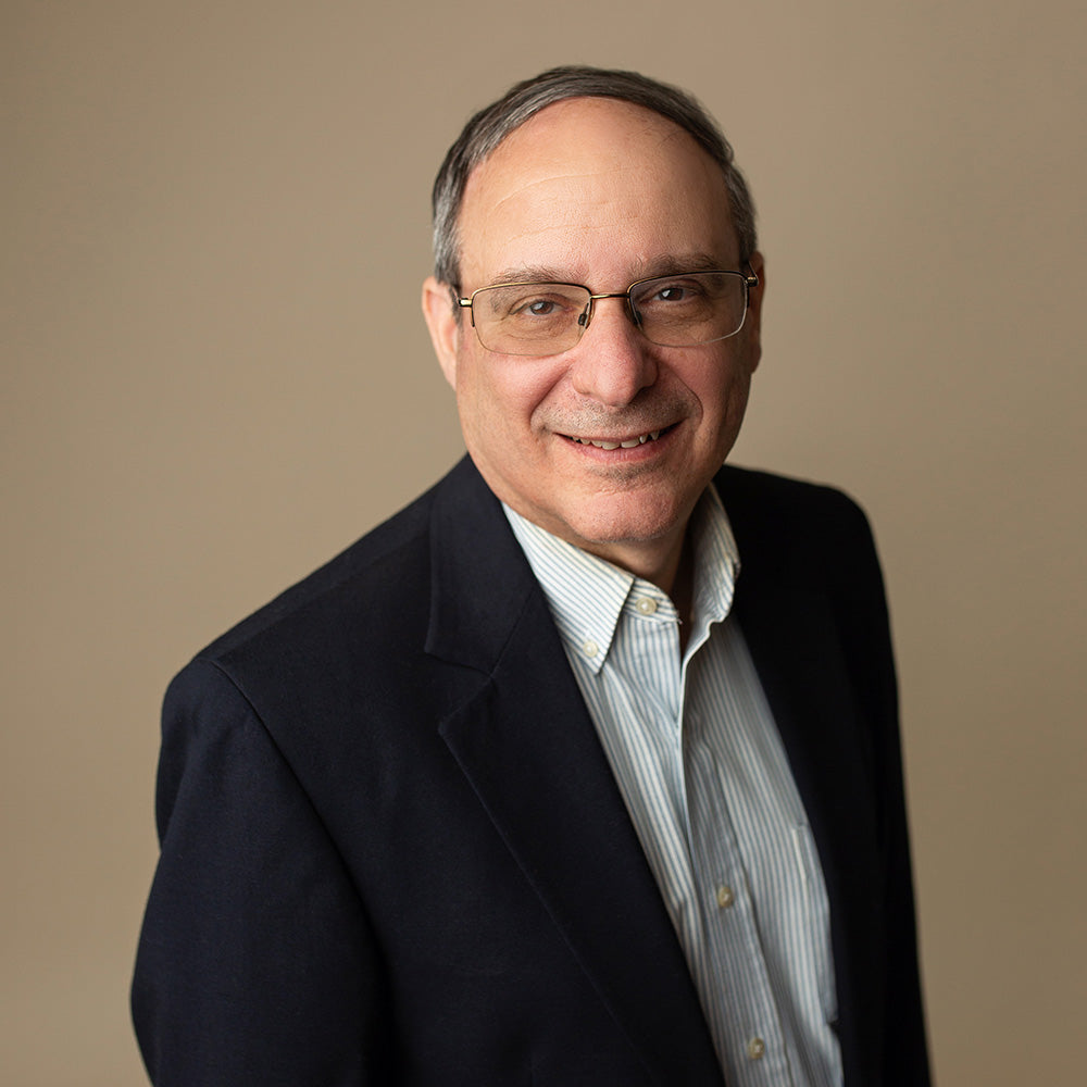 David Sandelman wearing glasses and a dark suit against a beige background