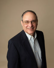 David Sandelman wearing glasses and a dark suit against a beige background