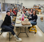 Group of people sitting around a table in a casual setting, with pizza boxes and drinks.